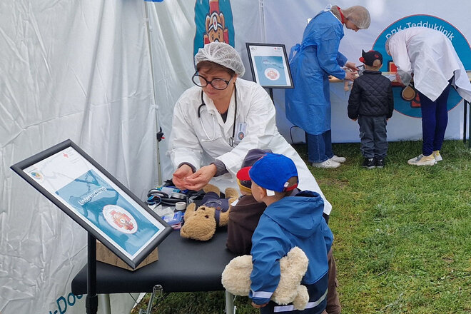 Eine Puppendoktorin mit großer Brille untersucht einen Teddybär, ein Kind schaut interessiert zu